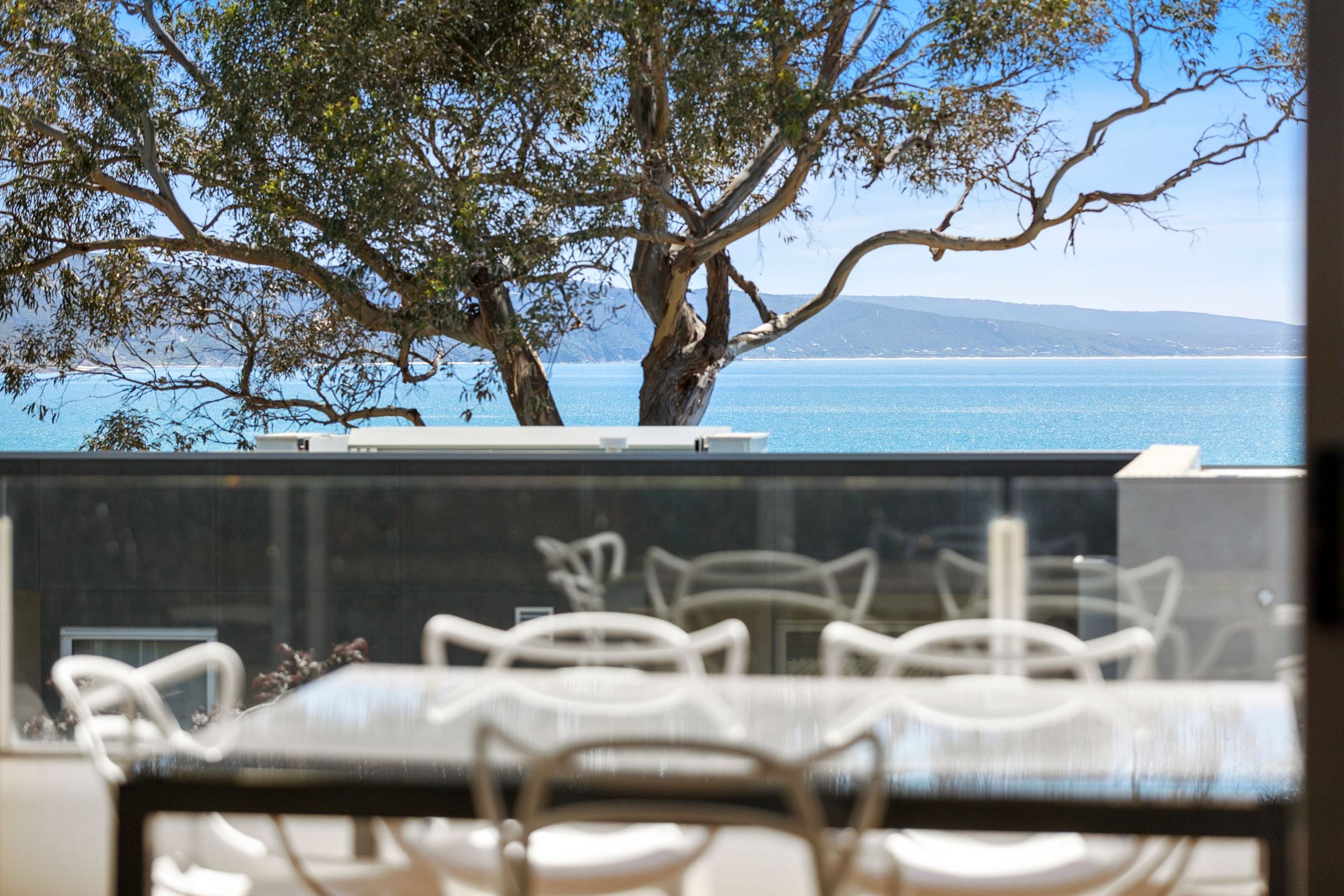 A dining area with white chairs and a table, overlooking a body of water and a tree.