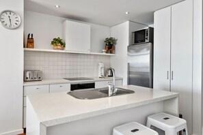 A modern kitchen with white cabinetry, a stainless steel refrigerator, a built-in oven, and a sink with a stainless steel faucet. - Apartment 18 Kalimna (Lorne)