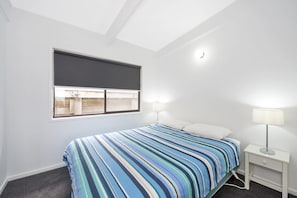 Ein Schlafzimmer mit einem Bett, einem Nachttisch, einer Lampe, einem Fenster mit Jalousie und einer Deckenleuchte.