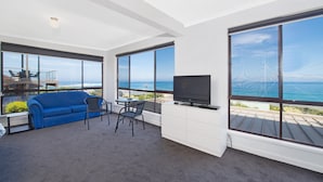 A modern living room with a blue sofa, a TV, and a dining area with chairs. Large windows offer a view of the ocean. - Cape Marengo (Marengo)