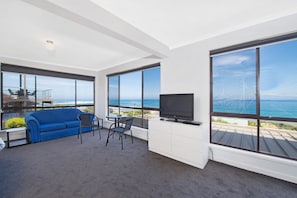 Ein modernes Wohnzimmer mit einem blauen Sofa, einem Fernseher und einem Essbereich mit Stühlen. Große Fenster bieten Blick auf den Ozean.
