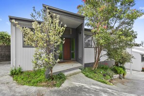 A modern two-story house with a front porch, a green door, and a small garden with flowering plants.