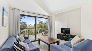 A modern living room with a sofa set, a coffee table, and a TV. There is a balcony with a wooden deck and a view of greenery. - Seabreeze - Central Lorne (Lorne)