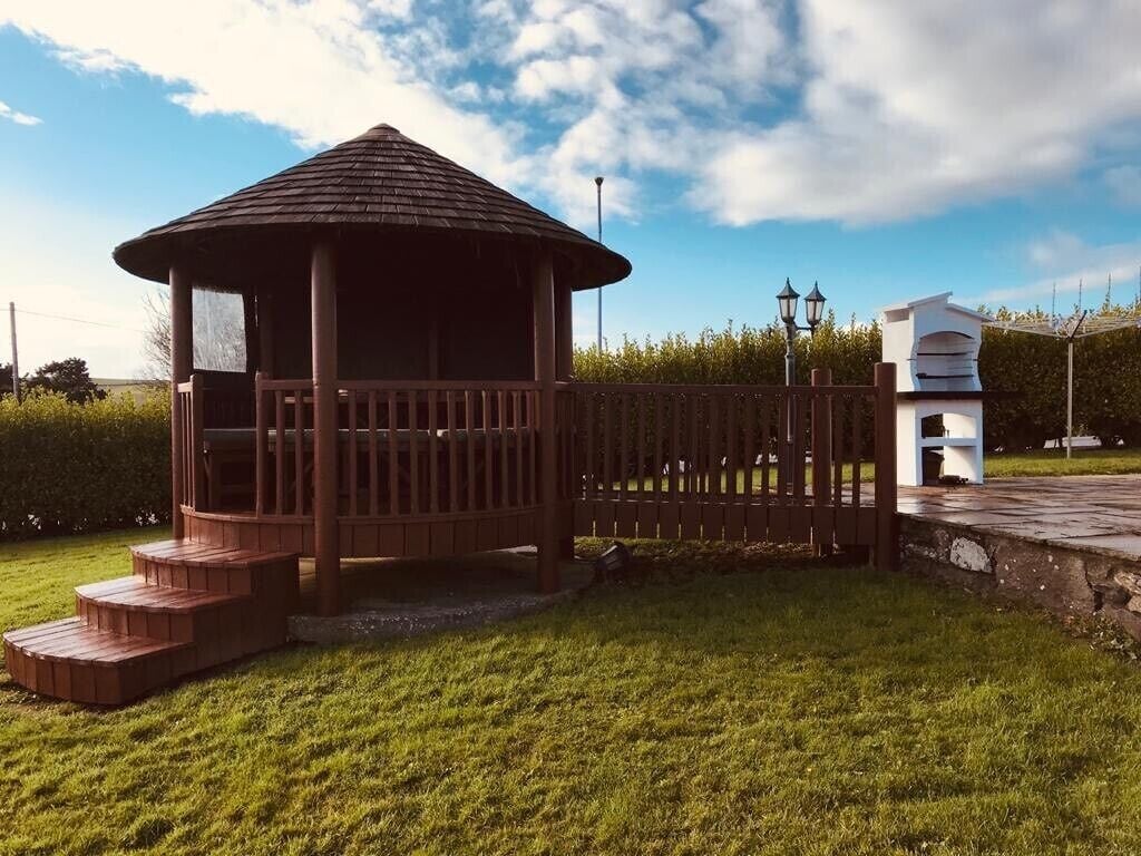 Un gazebo con struttura in legno e scalini, circondato da una recinzione e un edificio bianco sullo sfondo.
