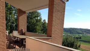 A covered patio area with a wooden table and chairs, brick pillars, and a view of a green landscape. - Fantastic house in Monferrato! (Tonco)