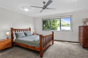Une chambre à coucher avec un lit en bois, une table de chevet, une commode, un ventilateur de plafond et une fenêtre donnant sur des arbres.