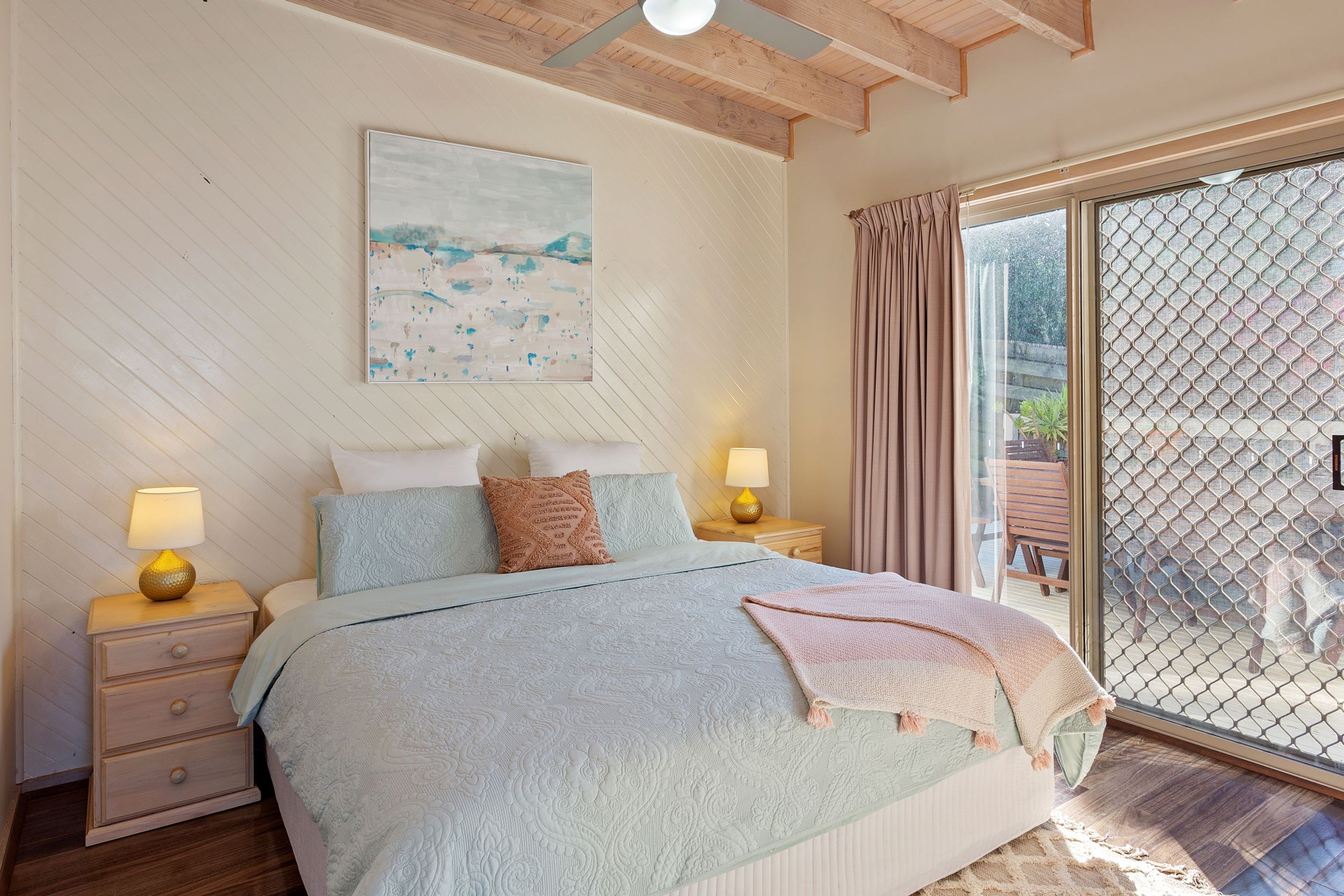 A bedroom with a bed, two bedside tables with lamps, a painting on the wall, and a view through a glass door.