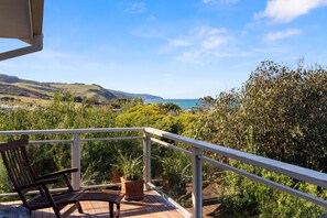 Ein Balkon mit einem Holzstuhl und einer Topfpflanze, mit Blick auf eine Küstenlandschaft mit Hügeln und dem Meer.