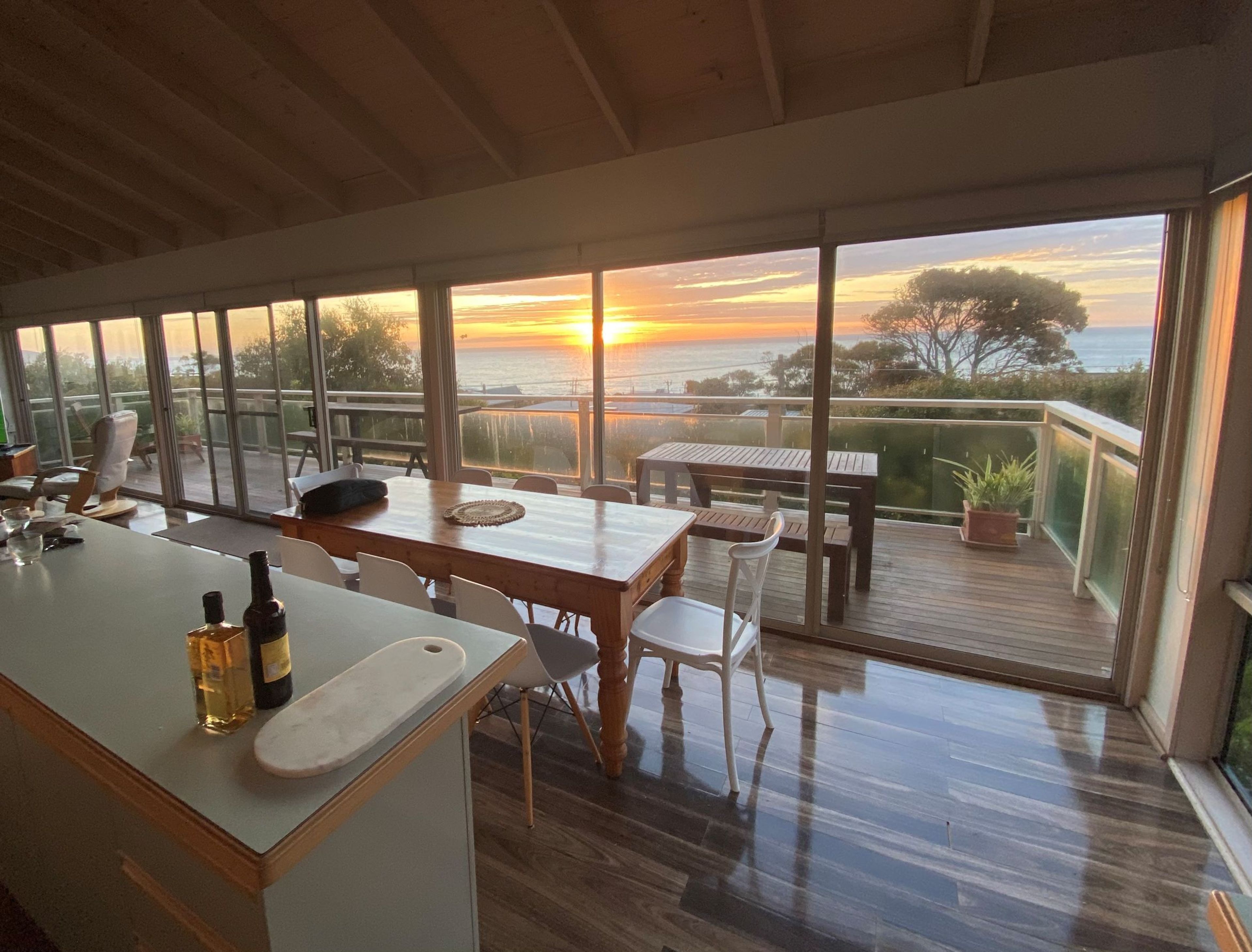 Ein Essbereich mit Blick auf den Sonnenuntergang über dem Meer, mit Holtmöbeln und einem verglasten Balkon.