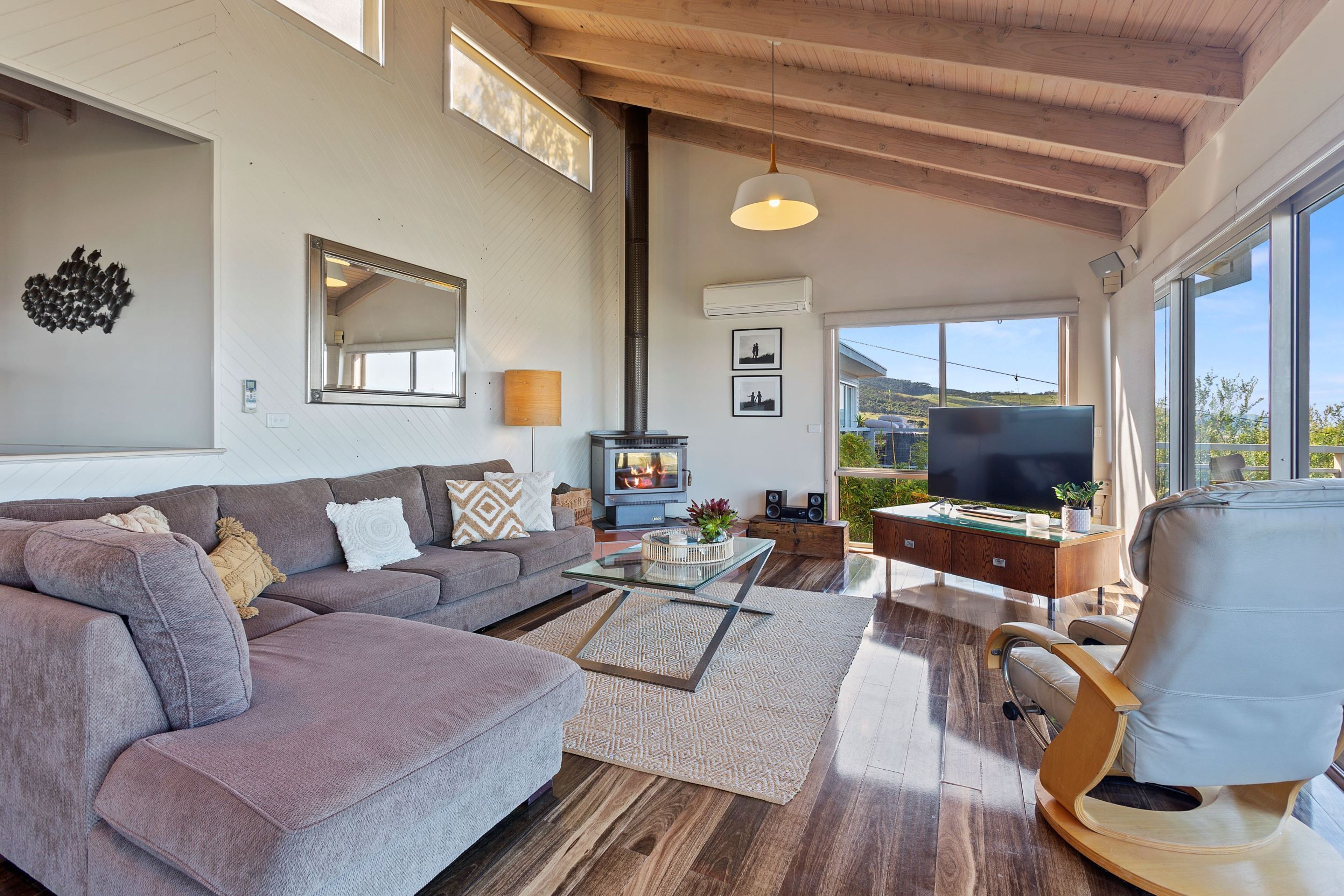 A spacious living room with a large sofa, a coffee table, and a television. There is a fireplace, a large window, and a ceiling with exposed beams.