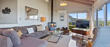 A spacious living room with a large sofa, a coffee table, and a television. There is a fireplace, a large window, and a ceiling with exposed beams.