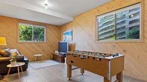 Ein gemütliches Wohnzimmer mit einem Kicker, einer Couch, einem Fernseher und großen Fenstern mit Blick auf Grünflächen.