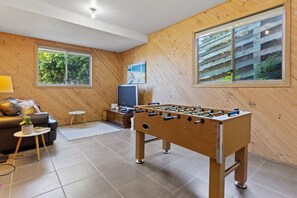 Ein gemütliches Wohnzimmer mit einem Kicker, einer Couch, einem Fernseher und großen Fenstern mit Blick auf Grünflächen.