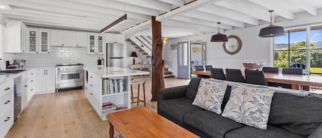 Ein großzügiger Wohnbereich mit einem Holztisch, einem grauen Sofa mit gemusterten Kissen und Blick ins Freie durch große Fenster.