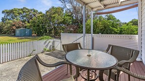 Eine überdachte Terrasse mit Korbmöbeln und einem Glastisch, umgeben von einem weißen Zaun und Bäumen.