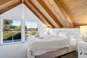 A bedroom with a sloped ceiling, large windows, a bed with white bedding, and a nightstand with a lamp.