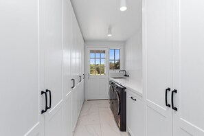 A narrow, white-walled laundry room with a built-in washer and dryer, a window with a view of greenery, and a door leading to another room.