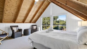 Ein Schlafzimmer mit schrägverdachter Decke, einem großen Bett, einem Sessel, einem kleinen Tisch und einem Fenster mit Blick ins Grüne.