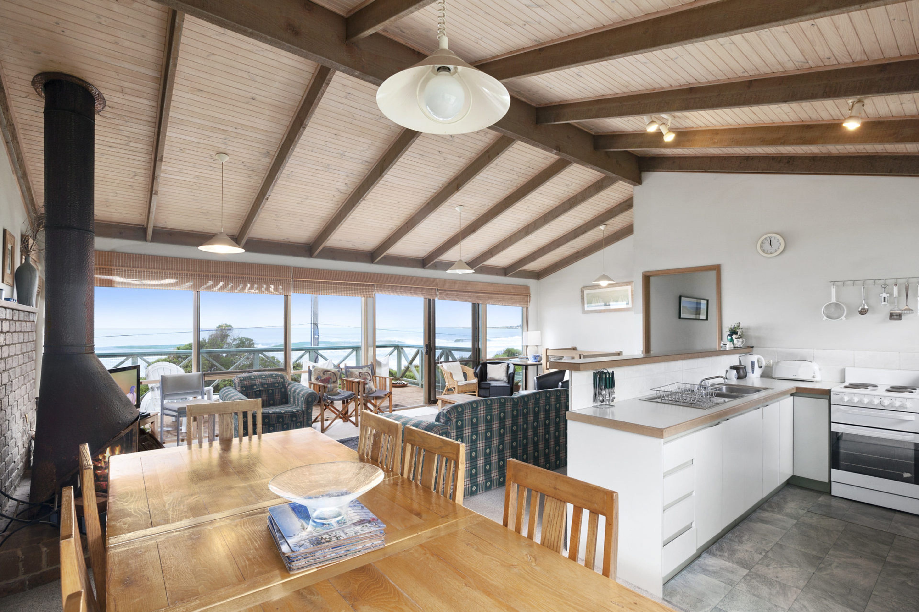 A spacious kitchen with a large island, wooden beams on the ceiling, and a view of the ocean through large windows.