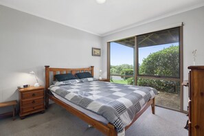 Ein Schlafzimmer mit einem hölzernen Bett, einem Nachttisch, einer Kommode und einem großen Fenster mit Blick auf Grünflächen.