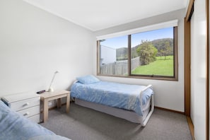 A bedroom with a single bed, a bedside table with a lamp, a dresser, and a window with a view of a green landscape.