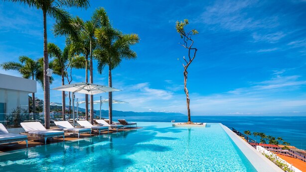 Piscina infinita com águas azul-claras, cercada por palmeiras e espreguiçadeiras, com vista para o oceano.