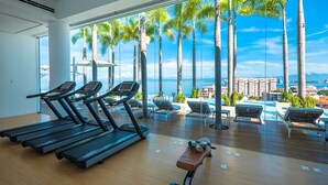 Academia com esteiras e um banco, com vista para uma piscina com espreguiçadeiras e palmeiras.