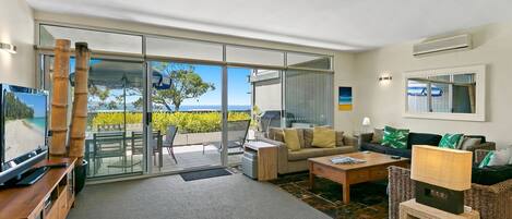 A spacious living area with a large sliding glass door leading to a balcony, a comfortable sofa set, a coffee table, and a television.