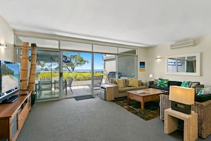 A spacious living area with a large sliding glass door leading to a balcony, a comfortable sofa set, a coffee table, and a television.