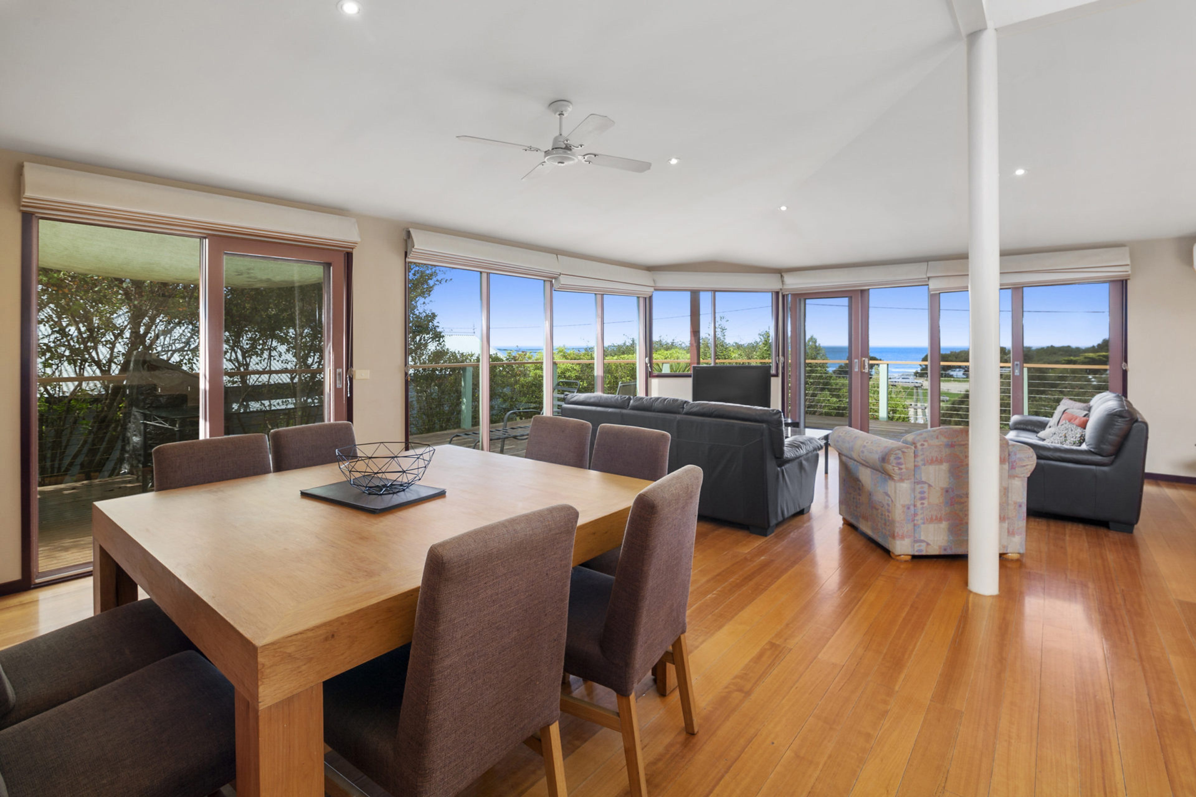 A spacious living area with a wooden dining table, multiple seating options, and large windows offering a view of the outdoors.