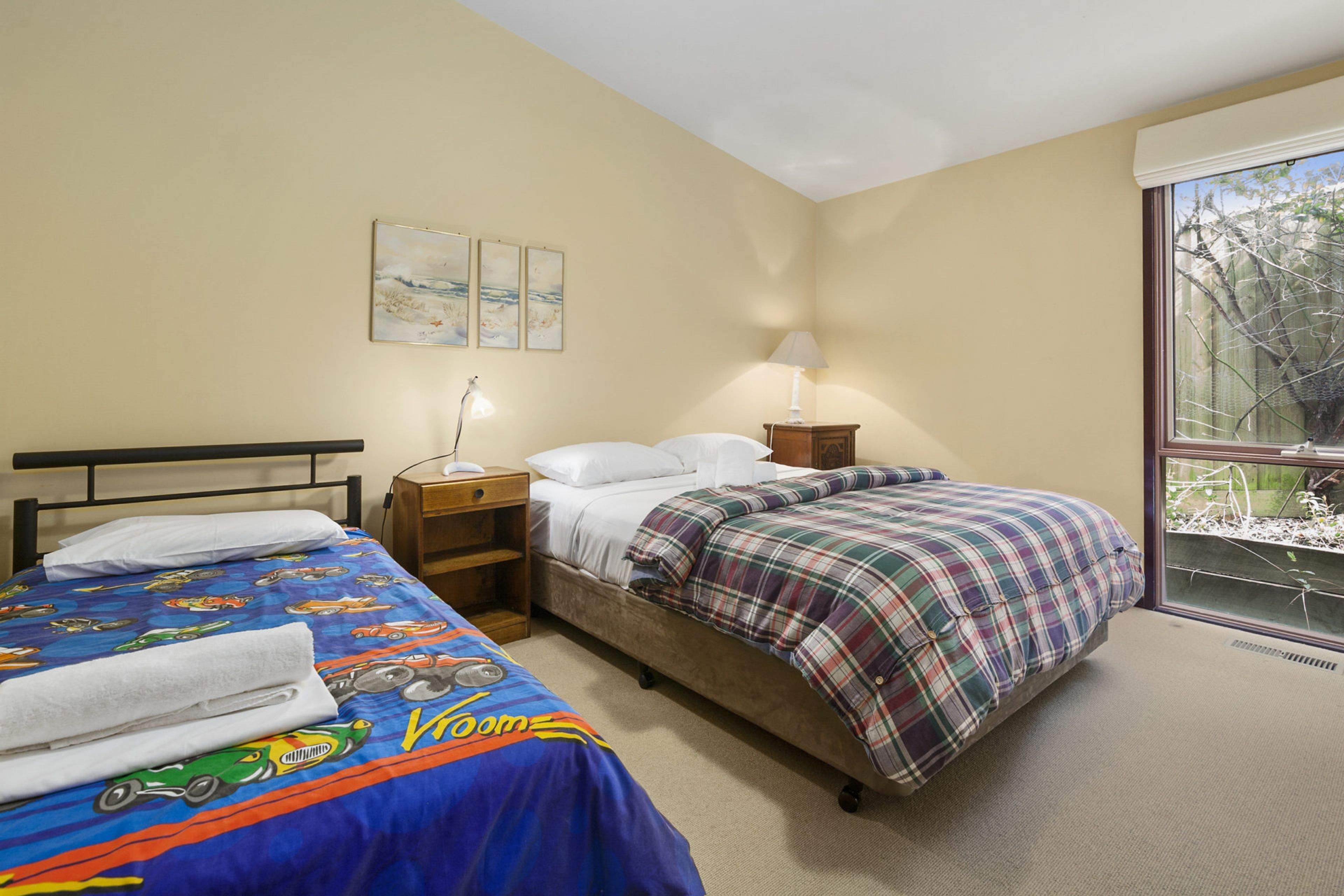 A bedroom with two beds, a nightstand, a lamp, and a window with a view of trees.