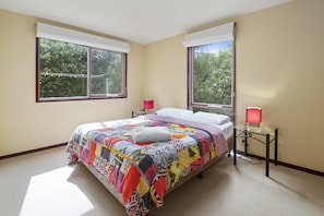 A bedroom with a large bed, two windows, a bedside table with a lamp, and a view of trees outside.
