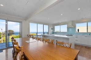 A modern kitchen with a large island, wooden dining table, and chairs, with a view of the sea through large windows. - Mirimar (Skenes Creek, Apollo Bay)