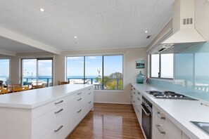A modern kitchen with a large island, built-in oven, and a view of the sea through large windows.