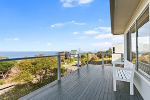 A wooden deck with a white bench, overlooking a coastal landscape with a clear sky.