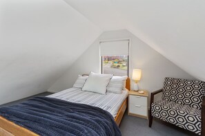 Ein Schlafzimmer mit schrägem Dach, einem Bett mit gestreifter Bettwäsche, einem Nachttisch mit Lampe und einem gemusterten Sessel.