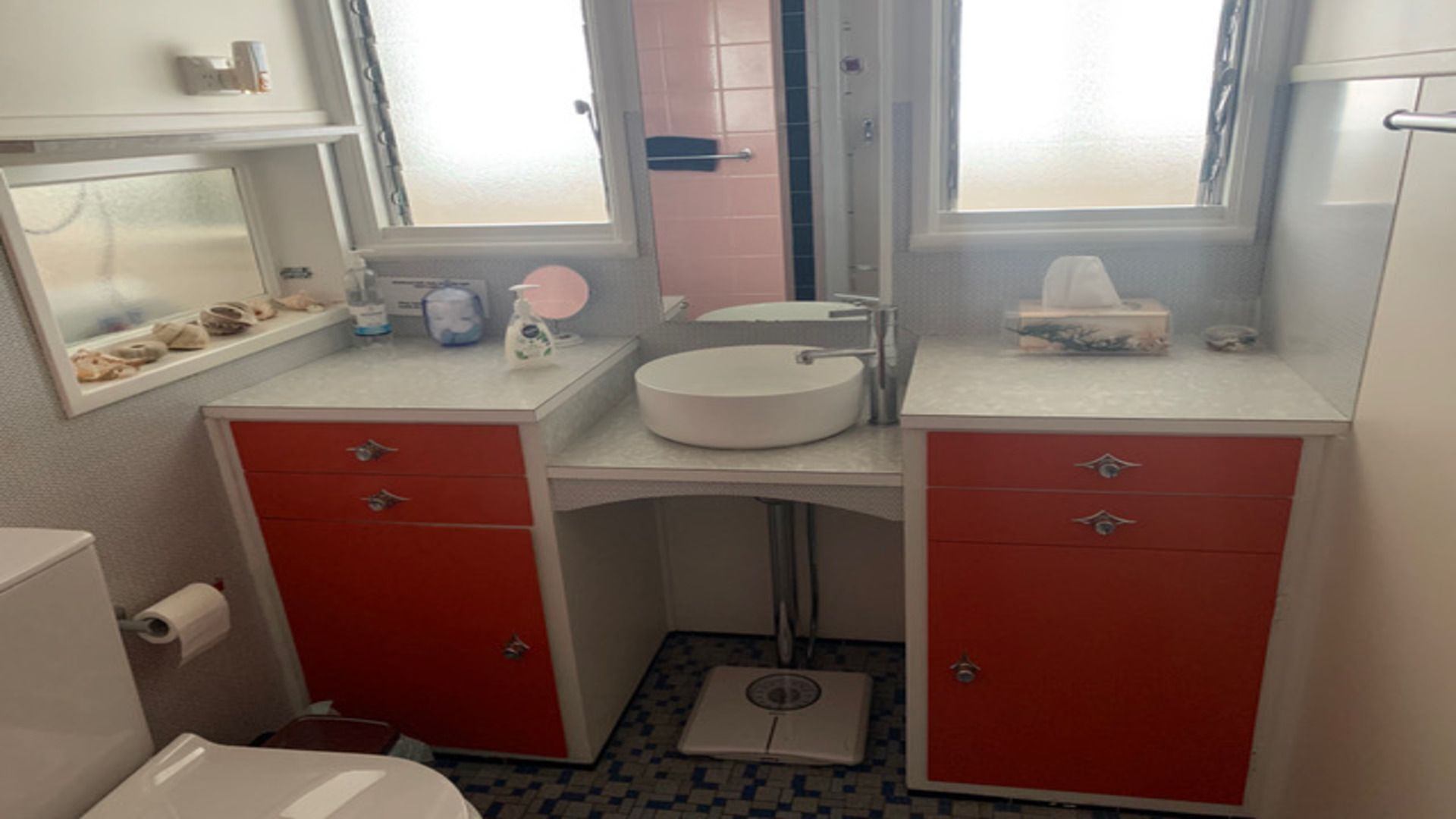Un baño pequeño con un lavamanos blanco, armarios rojos, una balanza y una ventana que deja entrar luz natural.