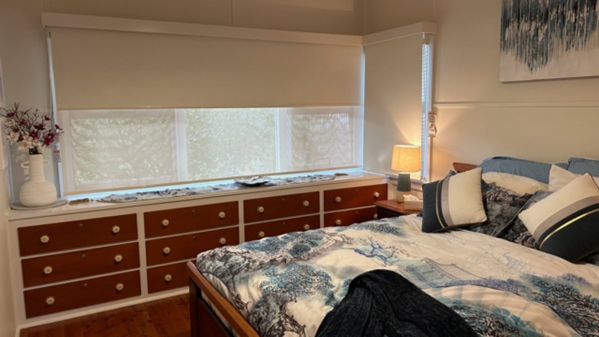 A bedroom with a large window, a dresser with multiple drawers, a bed with patterned bedding, and a bedside table with a lamp.