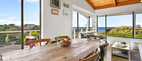 A dining area with a wooden table, chairs, and a bowl of fruit. There are large windows with a view of the sea and greenery.