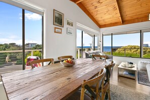 A dining area with a wooden table, chairs, and a bowl of fruit. There are large windows with a view of the sea and greenery. - Surf Reef (Marengo)