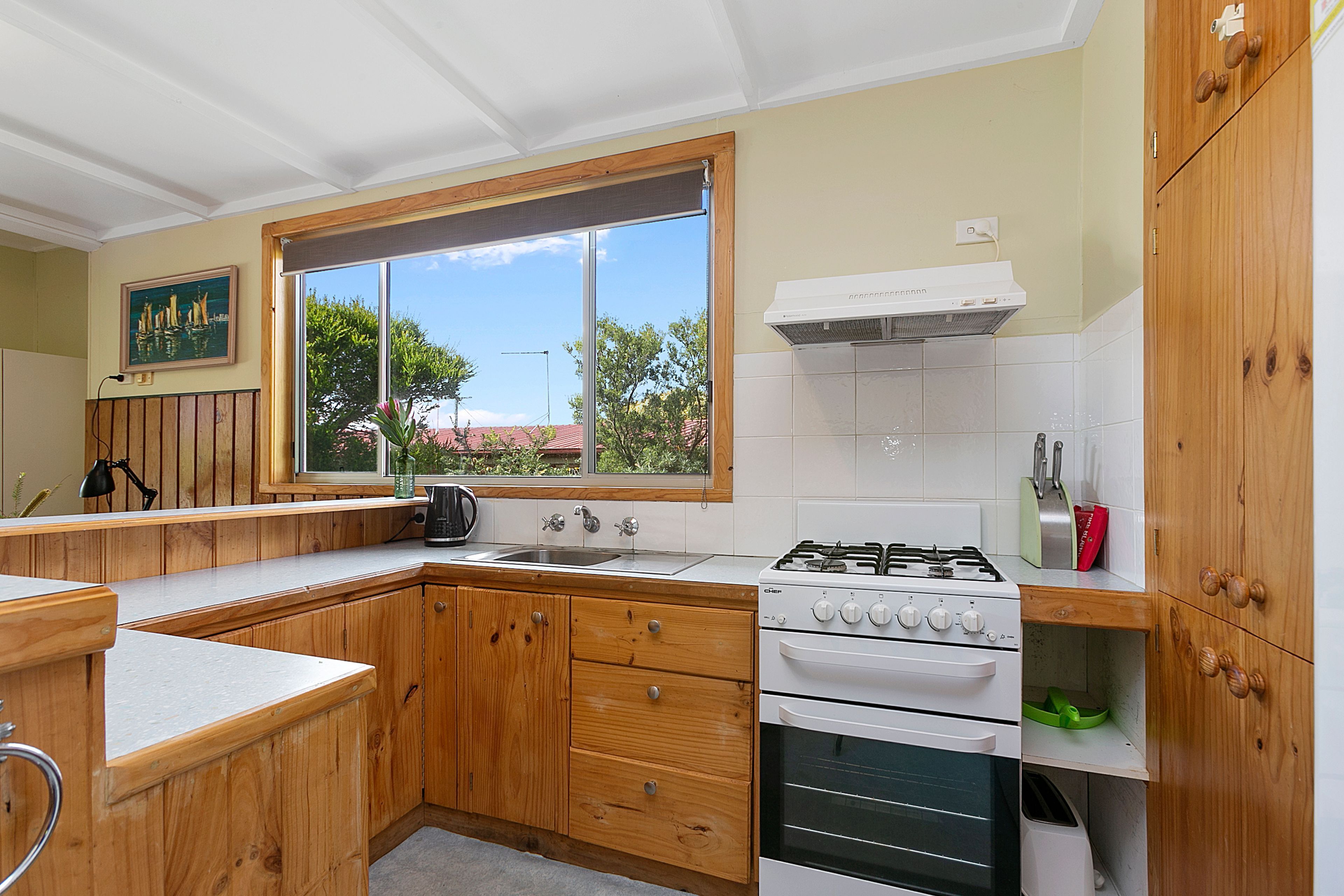 A kitchen with wooden cabinets, a gas stove, a sink, and a window with a view of trees.