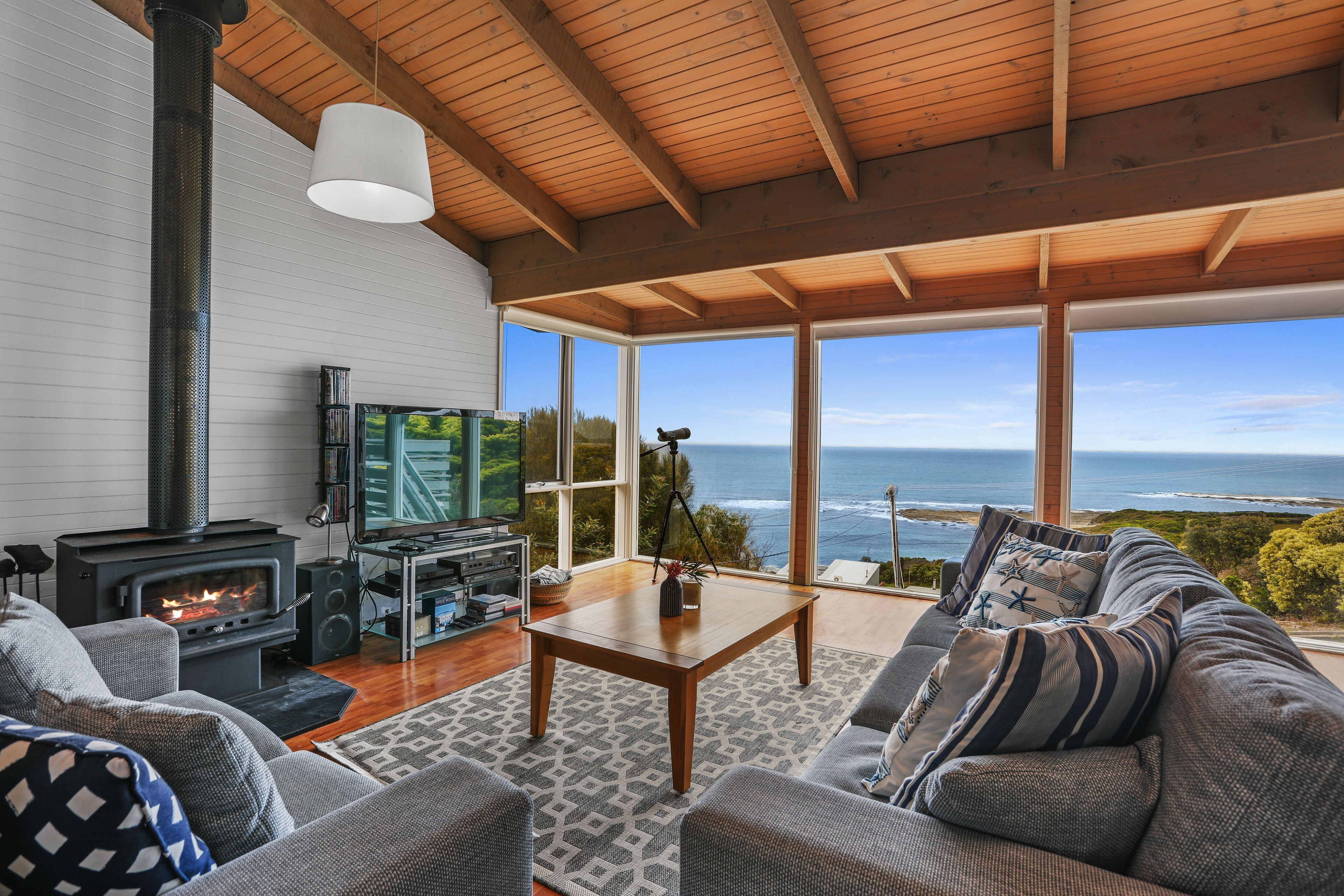 A cozy living room with a fireplace, a television, and a sofa facing a large window overlooking the ocean.