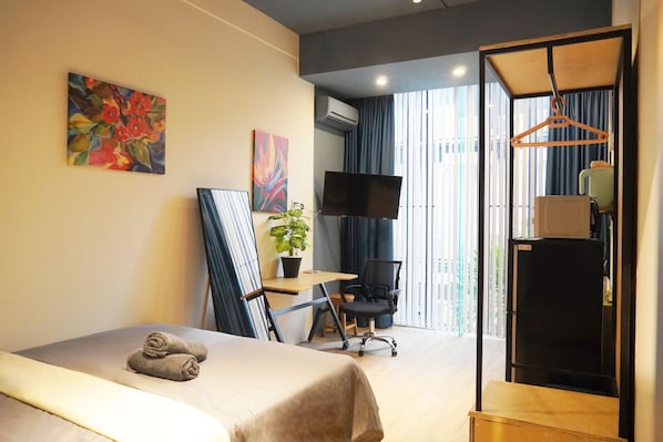 A hotel room with a bed, a desk, a chair, a TV, a plant, and a wardrobe.