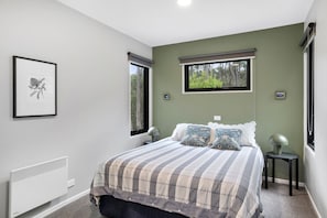A bedroom with a bed, two bedside tables, a radiator, and two windows with blinds.