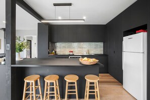 A modern kitchen with a central island, bar stools, dark cabinetry, and a white refrigerator.
