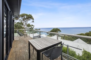 A wooden deck with a table and chairs, overlooking a body of water and greenery. - Summer By The Bay (Lorne)