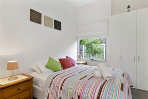 Ein Schlafzimmer mit Bett, Nachttisch, Lampe und einem Fenster mit Blick auf Grünflächen.