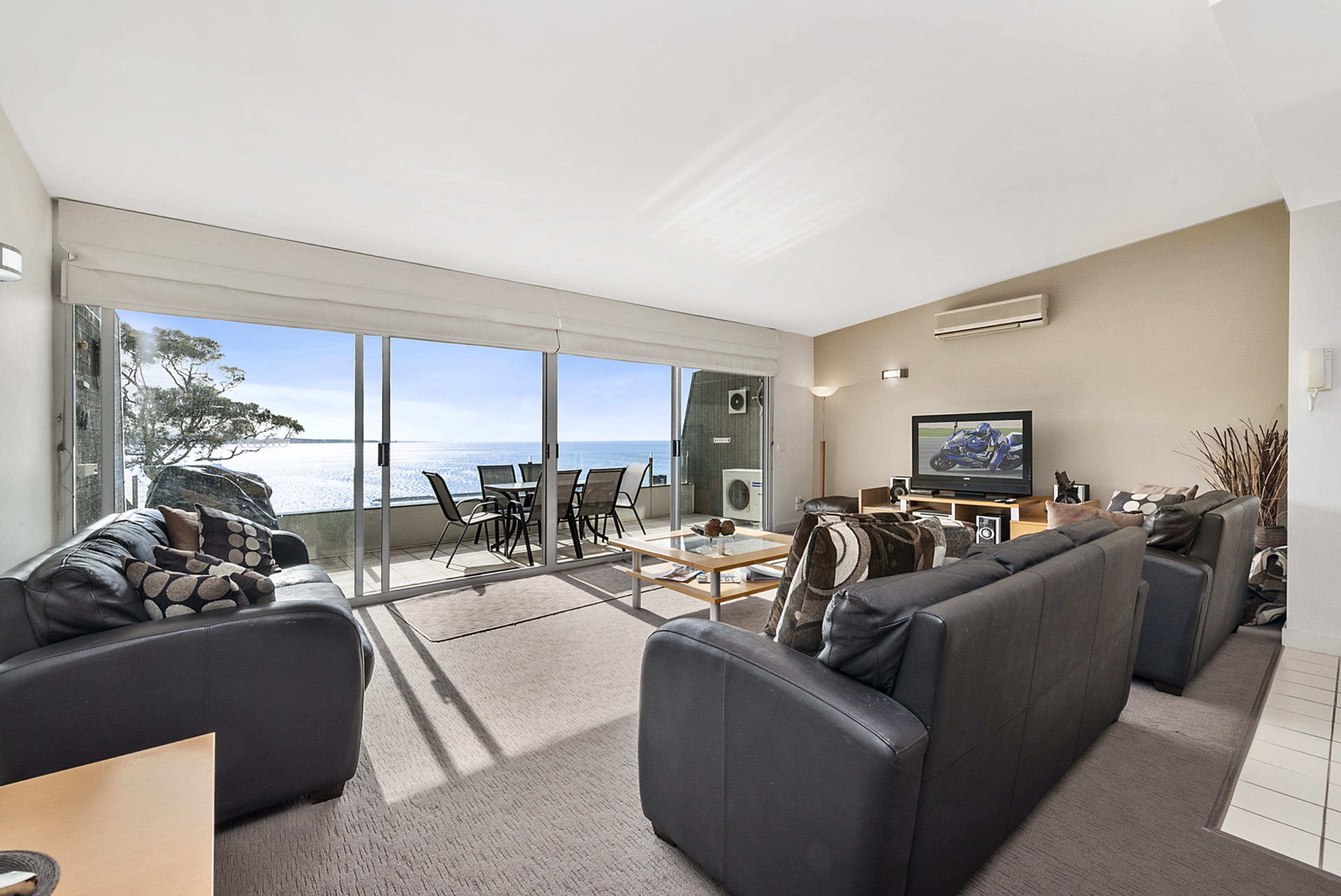 A modern living room with a large sliding glass door leading to a balcony with a view of the sea. There is a flat-screen TV mounted on the wall, a sofa, and a coffee table.