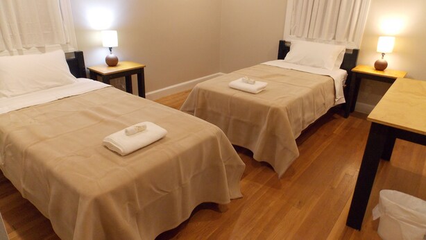 A hotel room with two beds, each with a beige bedspread and white pillows. There are two bedside tables with lamps, a wooden floor, and a small table with a vase. - The House of CROY (Alexandria)