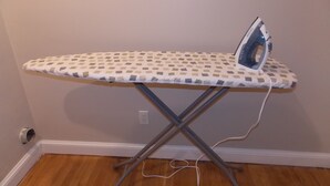 An ironing board with an iron on it, positioned against a wall with a wooden floor. - The House of CROY (Alexandria)